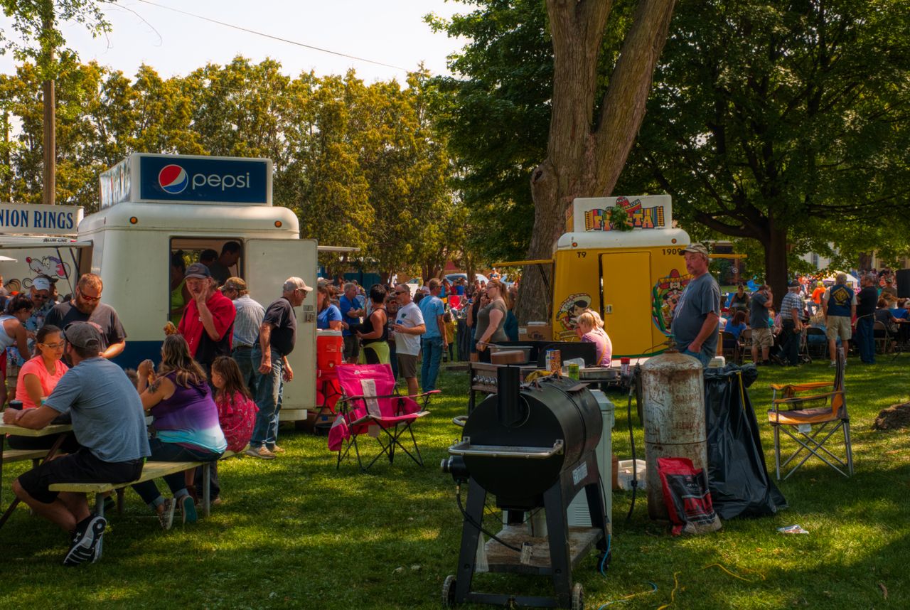 Crowd gathered around food trailers and a barrel smoker at a tailgate event